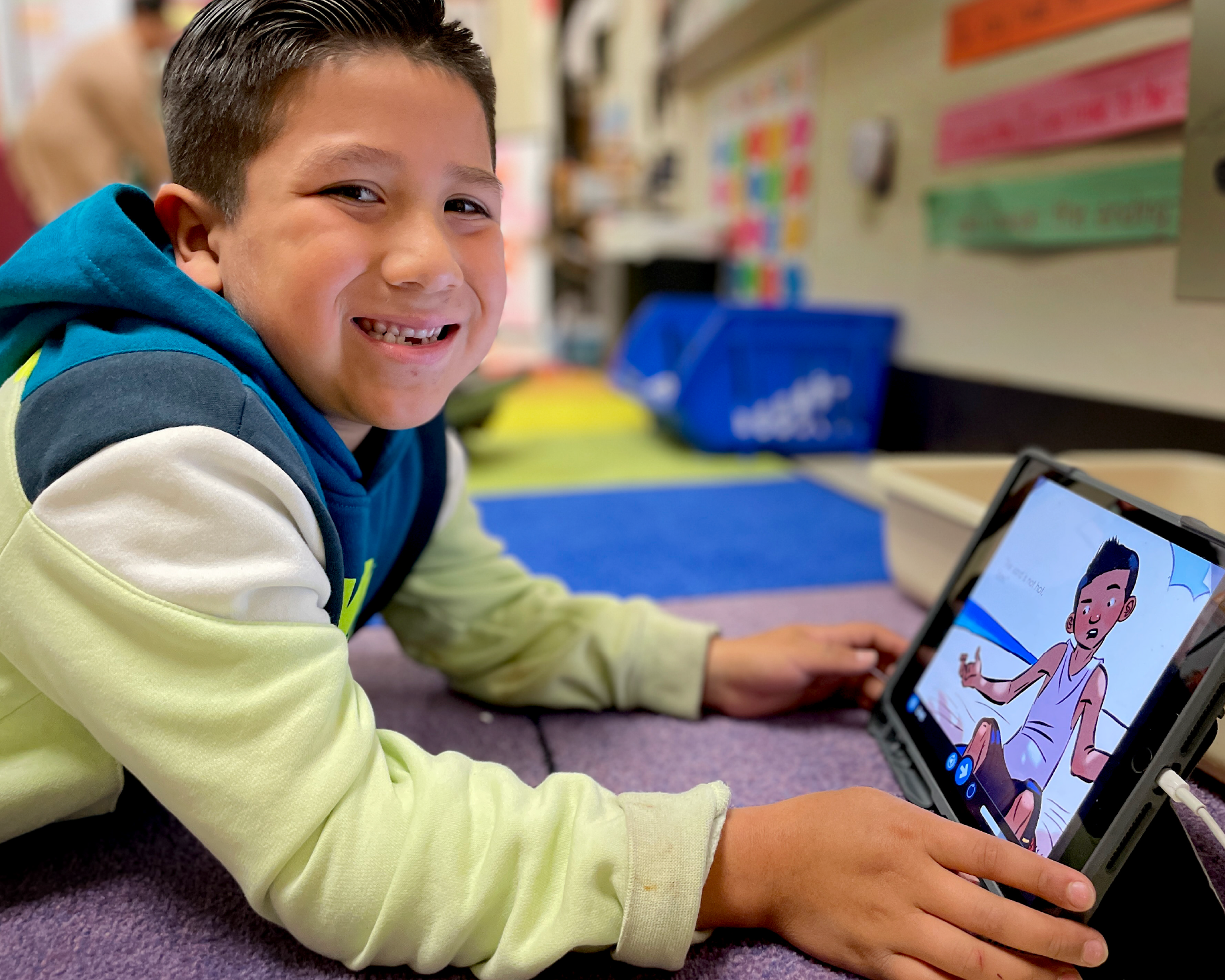 A young boy with a bright smile is sitting on the floor and using a tablet device, surrounded by a colorful classroom environment with posters and other educational materials on the walls.