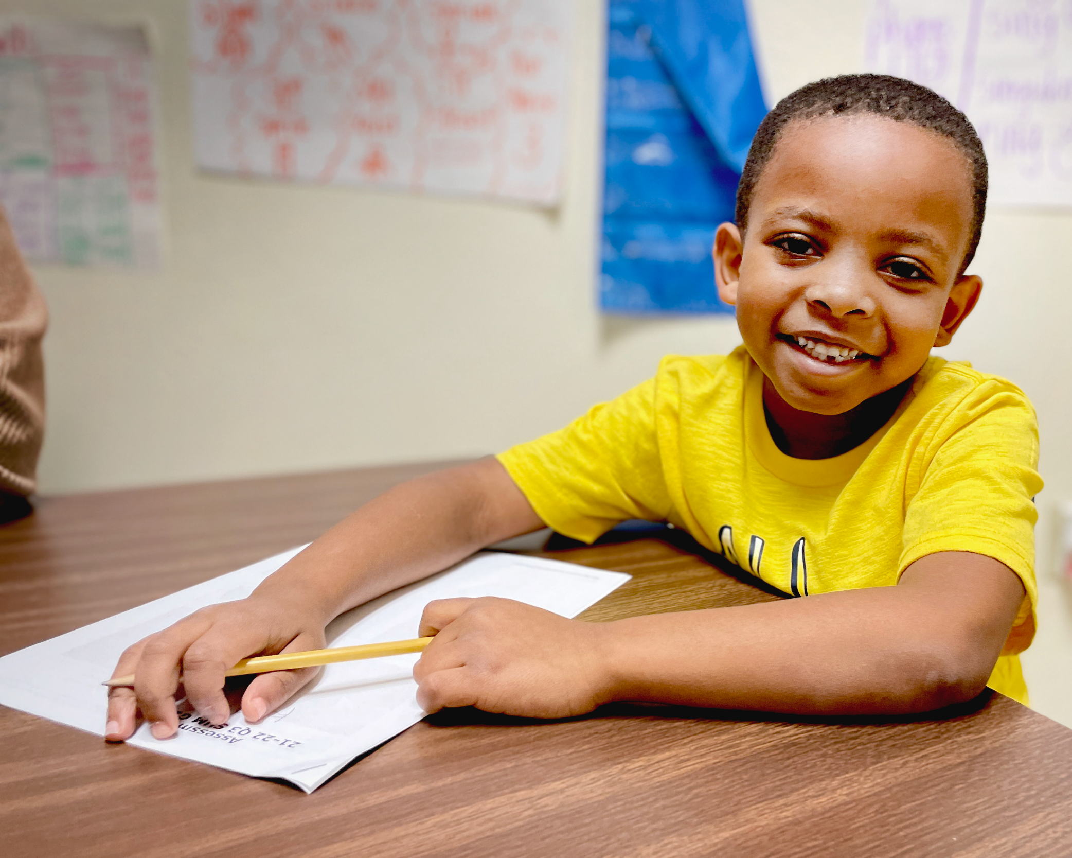 A young boy in a yellow shirt is sitting at a desk, smiling and drawing on a piece of paper. The background includes colorful artwork and posters on the walls, suggesting an educational or classroom setting.