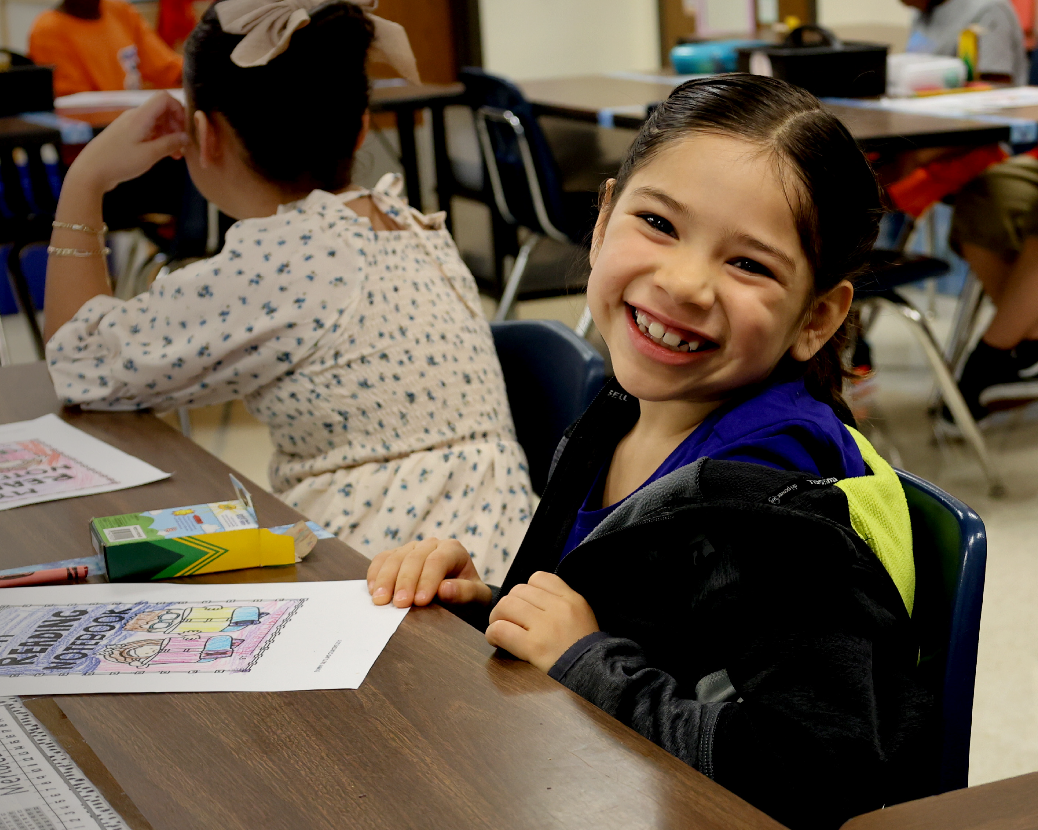 A young girl in a school setting, smiling brightly while working on an activity at her desk, with other students visible in the background.