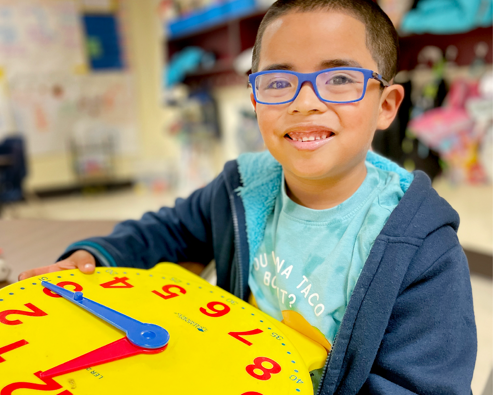 A young boy with glasses is smiling while holding a colorful clock toy in what appears to be a classroom or educational setting.
