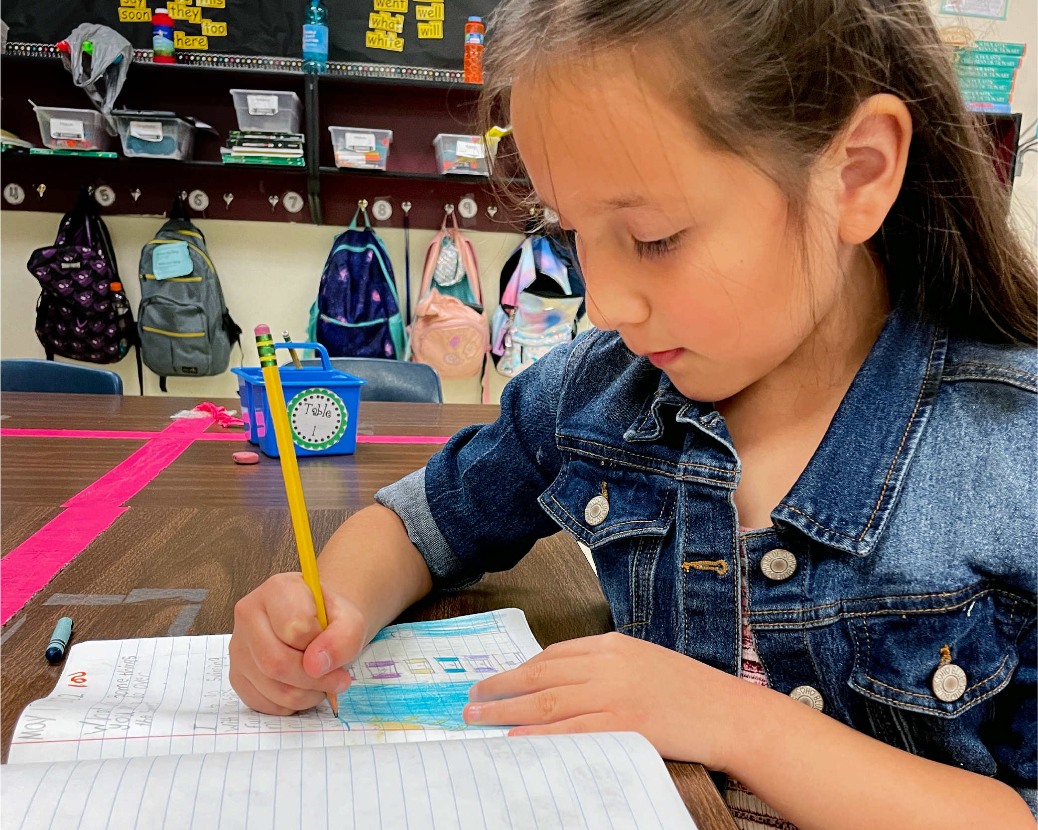A young girl in a denim jacket is intently focused on writing in a notebook on a desk, surrounded by colorful backpacks and school supplies in a classroom setting.