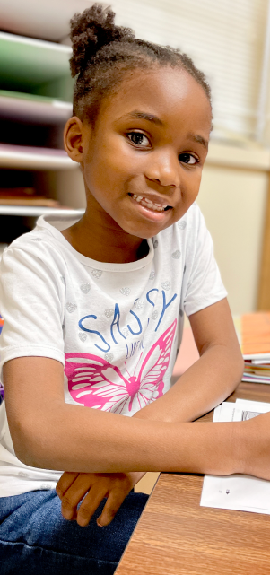 A young girl with braided hair wearing a white t-shirt with a butterfly design is sitting at a desk in what appears to be a classroom or educational setting.