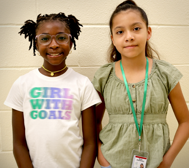 Two young girls, one wearing a "Girl with Goals" t-shirt and the other in a green dress, stand together in what appears to be a school or educational setting, with a plain white wall in the background.