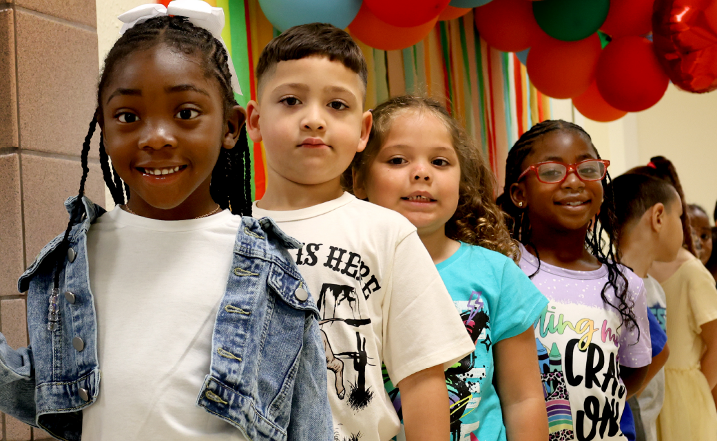 A group of diverse children, with different ethnicities and ages, are standing together in a colorful, festive environment with balloons and decorations in the background.