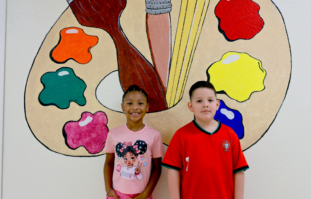 Two children, a girl and a boy, are standing in front of a colorful abstract painting with various shapes and patterns.