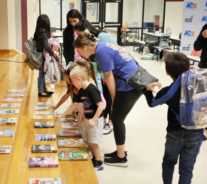 A group of people, likely students, are gathered around a table examining various books and materials in what appears to be a classroom or library setting.