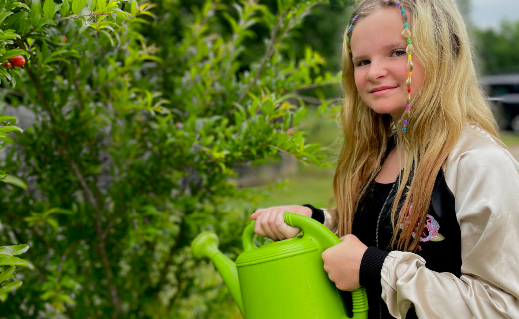 A young woman with long blonde hair stands in a lush, green garden, holding a green watering can and smiling at the camera.