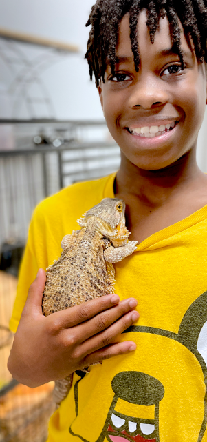A young person with a warm smile is holding a small lizard-like creature in their hands, with a wooden structure visible in the background.