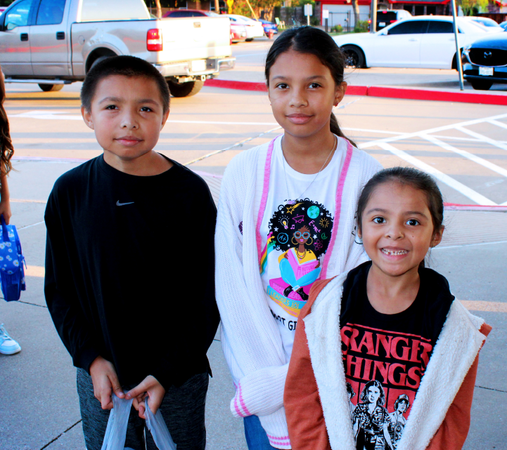 Three young children, two girls and one boy, are standing together on a city street. In the background, there are various vehicles and buildings visible.