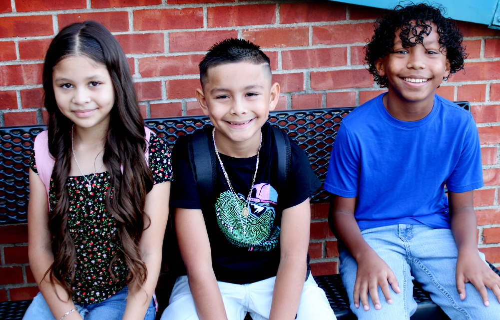 Three young individuals, a girl and two boys, are standing in front of a red brick wall, appearing to be smiling and posing for the camera.