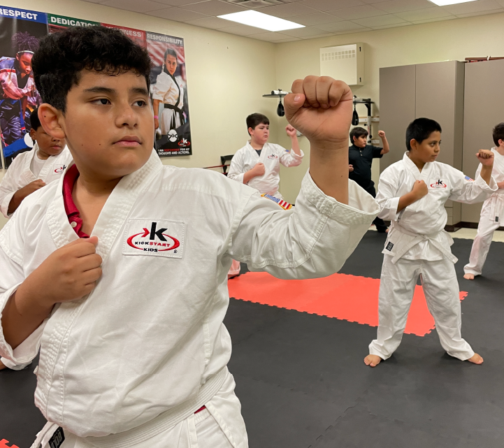 The image shows a group of young individuals in white martial arts uniforms practicing in a martial arts studio, with one person in the foreground raising their fist in a determined stance.