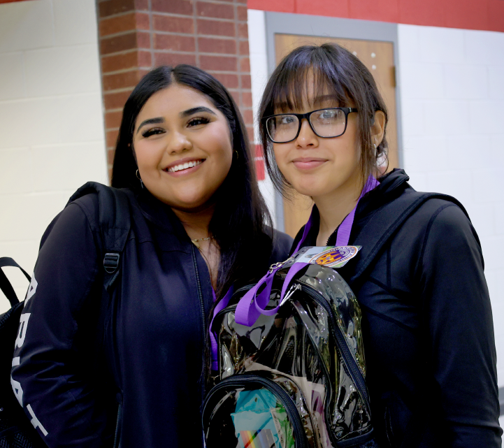 Two young women, one with glasses and the other with a bright smile, stand together in front of a brick building.