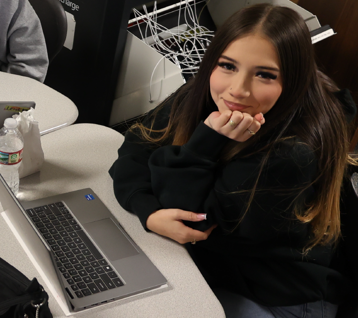A young woman with long dark hair is sitting at a desk, using a laptop computer in what appears to be an office or workspace environment.