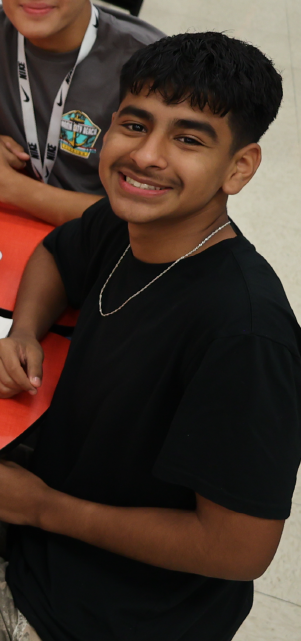 A young boy with a warm smile, wearing a black shirt, sits in what appears to be a classroom or educational setting.