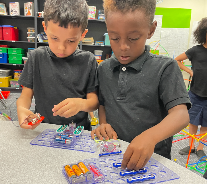 Two young boys, one with short dark hair and the other with curly hair, are intently focused on playing with colorful toys and objects on a table in what appears to be a classroom or educational setting, with shelves and storage containers visible in the background.