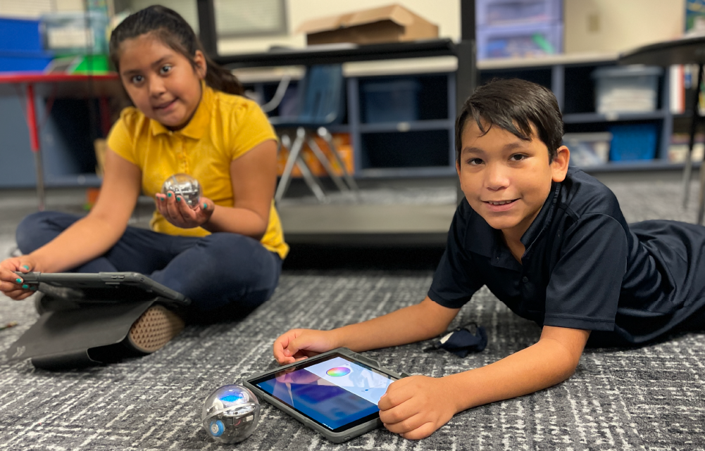 Two young students, a girl in a yellow shirt and a boy in a black shirt, are sitting on the floor and using a tablet device together in what appears to be a classroom setting with bookshelves and other educational materials visible in the background.
