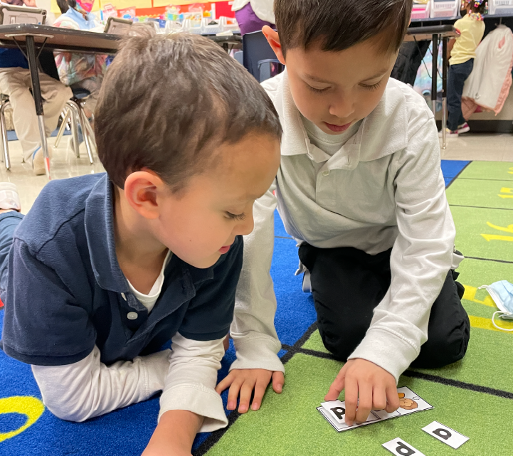Two young boys sitting on the floor, engaged in an educational activity with cards or letters in a classroom setting with colorful decorations on the walls.