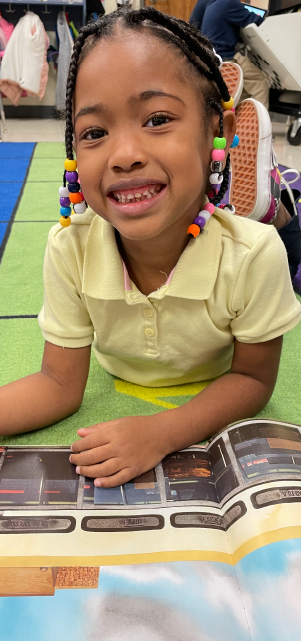 A young girl with braided hair and a bright smile is sitting on the floor, surrounded by colorful educational materials and toys.