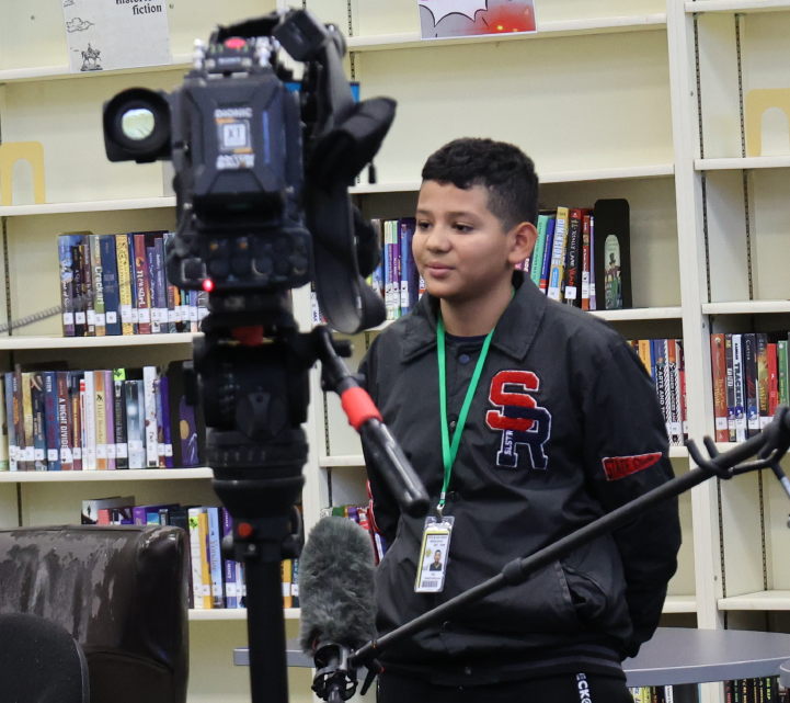 A young boy wearing a black shirt with a logo stands in front of a camera setup, surrounded by bookshelves in the background.