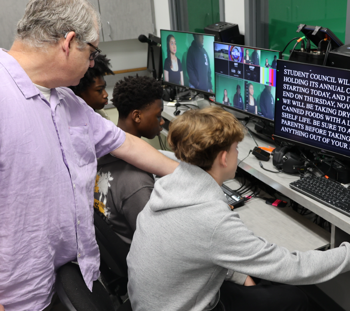 The image shows a group of students working together on computers, with a teacher or instructor assisting them. The background displays a screen with various visual elements, suggesting an educational or training setting.