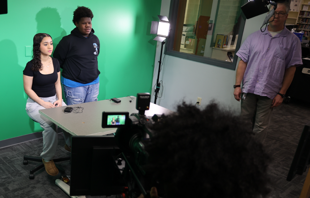 The image shows a group of people in a studio setting, with a green screen backdrop and various equipment and props visible in the foreground.