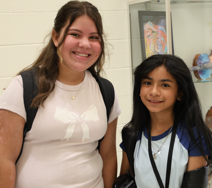 Two young women with friendly expressions are standing together in what appears to be a school or educational setting, with artwork visible in the background.