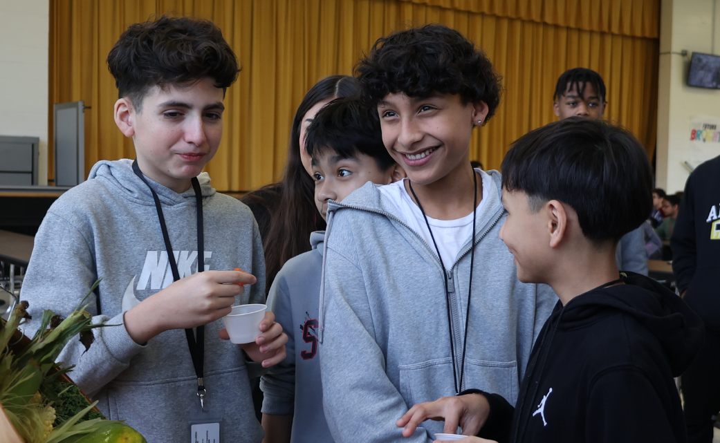 A group of young people, likely students, are gathered together and engaged in conversation, with a wooden wall and door visible in the background.