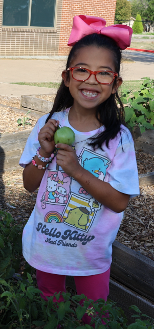 A young girl with a bright pink bow in her hair is smiling and holding a green apple, standing in front of a brick building with windows.