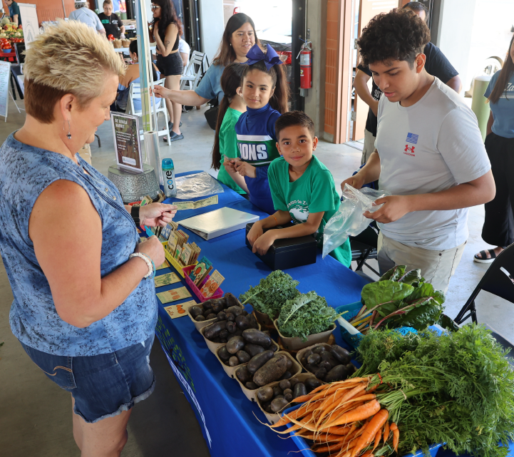A group of people, including children, gathered around a table displaying various fresh produce such as carrots, leafy greens, and other vegetables.