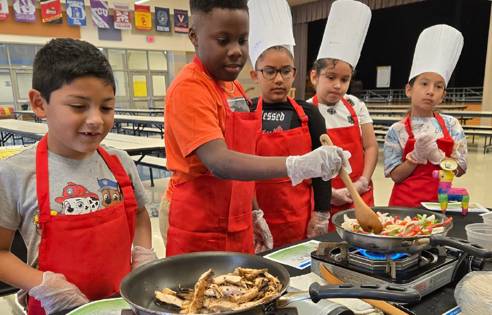 A group of young children, wearing red aprons, are gathered around a stove, cooking and preparing food together in what appears to be a classroom or educational setting.