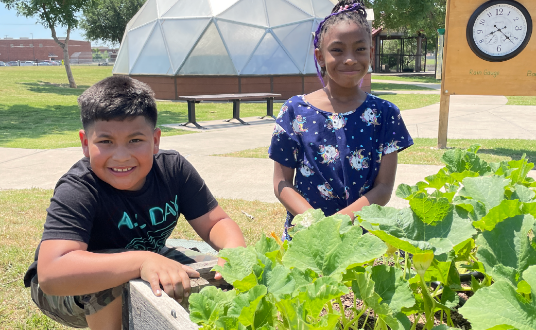Two young children, a boy and a girl, are standing in a garden setting surrounded by lush greenery and a geodesic dome structure in the background.