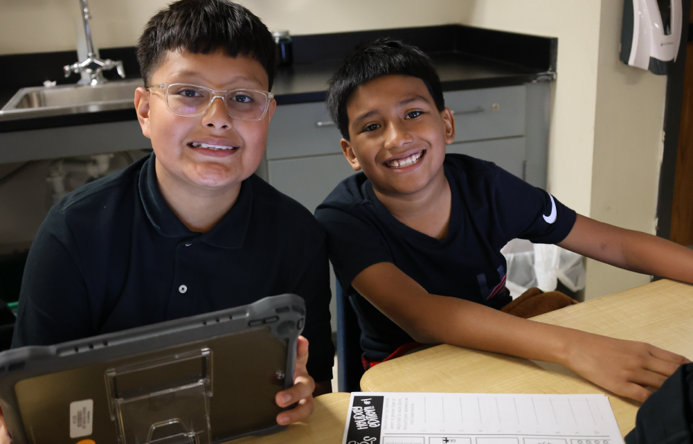 Two young individuals, one wearing glasses, are smiling and posing together in what appears to be a kitchen or work environment.