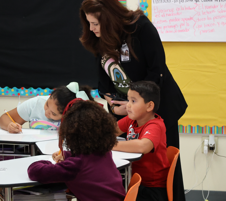 The image shows a teacher assisting students in a classroom setting, with colorful decorations and materials visible in the background.