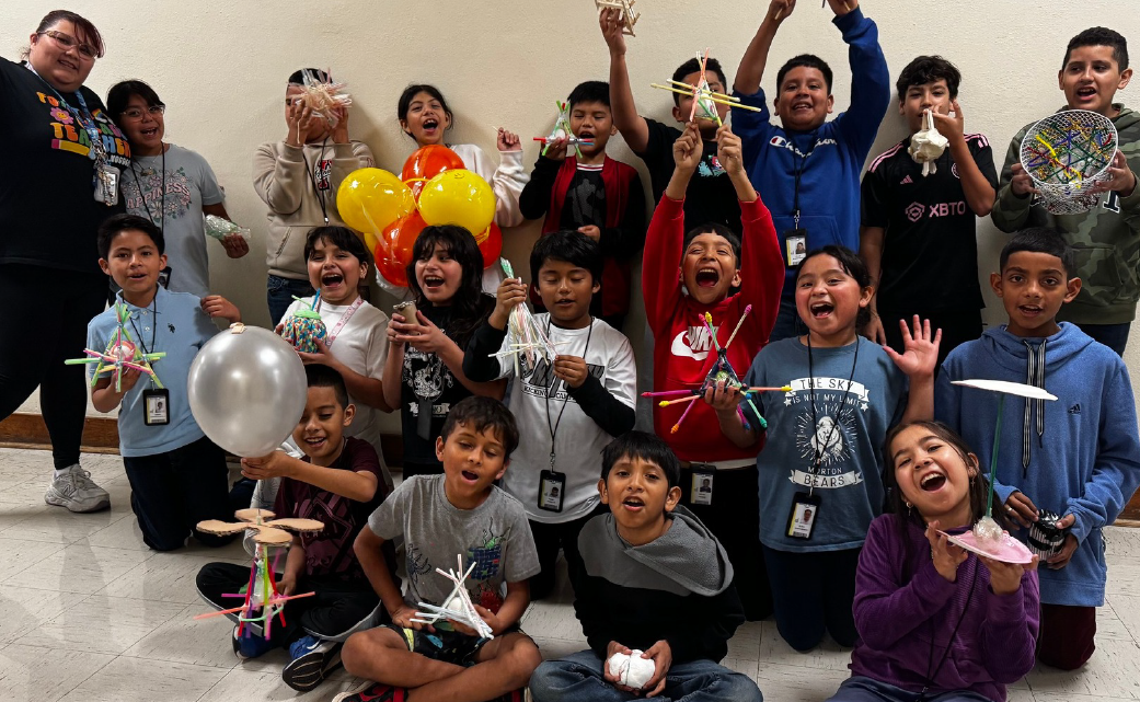 A group of enthusiastic children, some wearing colorful clothing and holding various props, appear to be engaged in a lively activity or celebration in what seems to be an indoor setting with a plain white wall in the background.