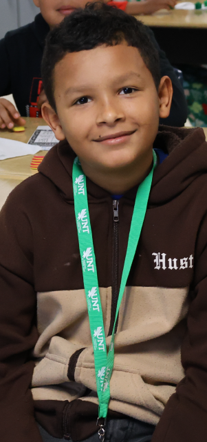 A young boy wearing a brown hooded jacket and a green lanyard is smiling at the camera in what appears to be a classroom or educational setting.