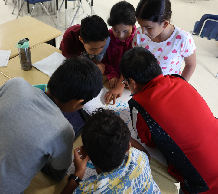 A group of young students, both boys and girls, are gathered around a table, intently focused on a task or activity they are engaged in together.