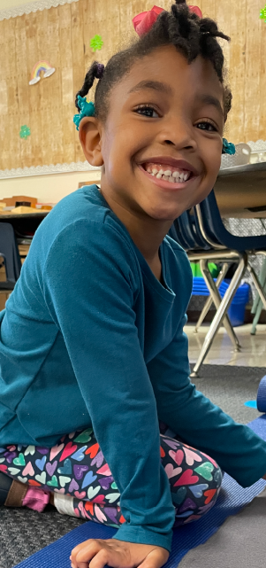 A young girl with a bright smile, wearing a teal shirt and patterned pants, sitting in a classroom setting with colorful decorations in the background.