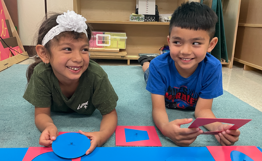 Two young children, a girl and a boy, are sitting on the floor in a classroom setting, playing with colorful educational toys and smiling happily.