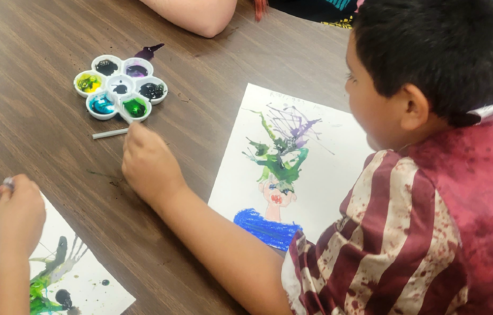 A young child is sitting on the floor, painting a colorful abstract artwork on a canvas using various paints and brushes.