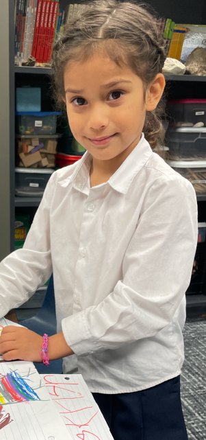 A young girl with curly hair is sitting at a desk, surrounded by shelves and drawers in the background.