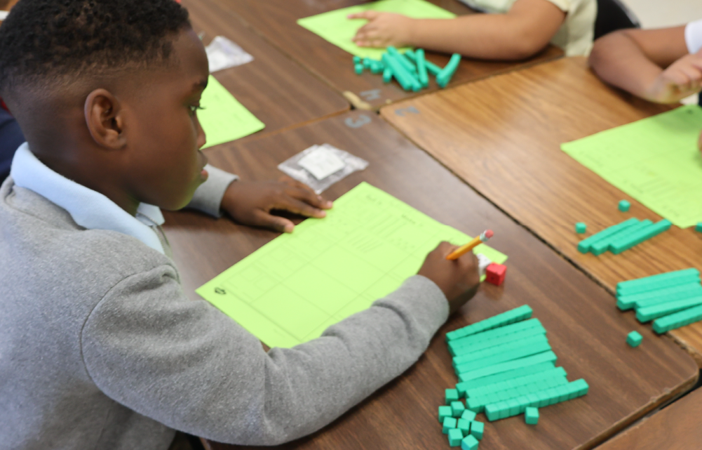 A young boy in a gray sweater is sitting at a table, intently working on a craft project with colorful paper and materials.