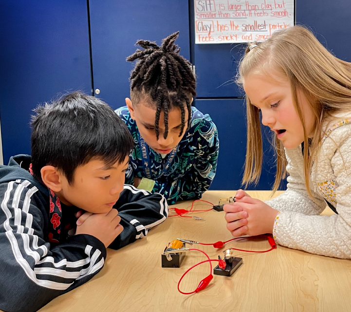Three children, two boys and one girl, are engaged in a hands-on learning activity, working with electronic components on a table in a classroom setting.