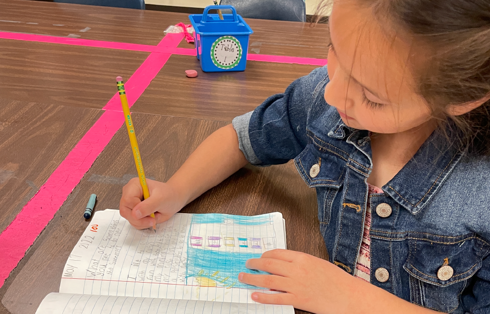 A young girl in a denim jacket is sitting on the floor, intently focused on writing in a notebook with a colorful pen, surrounded by a blue toy radio and pink tape on the wooden floor.
