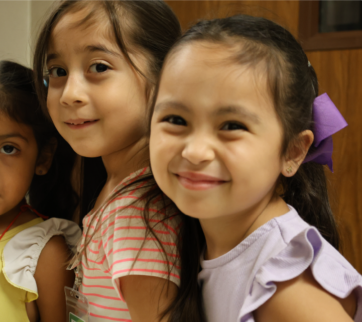 The image shows two young girls with warm, friendly expressions, standing together in what appears to be a home setting.