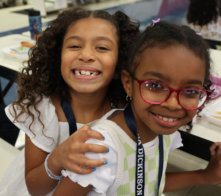 Two young girls with curly hair and bright smiles, one wearing glasses, appear to be in a classroom or educational setting.