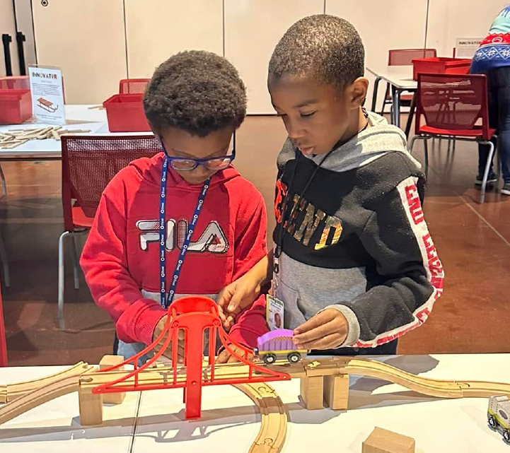 Two young boys, one wearing a red Fila shirt and the other a gray sweatshirt, are intently focused on playing with a toy train set on a table in what appears to be a classroom or educational setting, with various supplies and materials visible in the background.