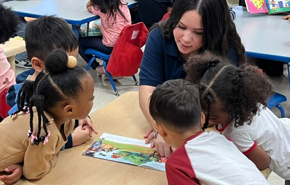 A group of young students, both boys and girls, are gathered around a teacher who is reading a book to them in a classroom setting.