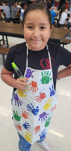 A young child wearing a colorful apron with handprints, smiling brightly while holding a paintbrush in a classroom setting.