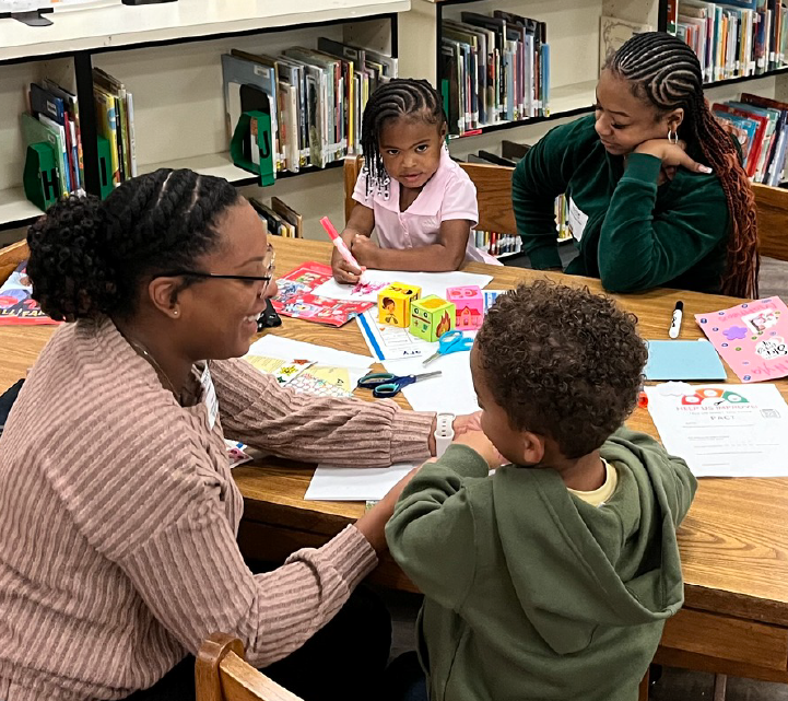 The image shows a group of young students working together at a table in what appears to be a classroom or library setting, surrounded by bookshelves in the background.