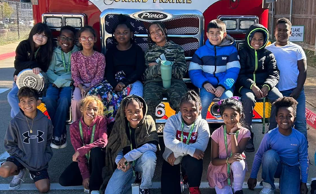 A group of diverse individuals, both adults and children, are gathered in front of a fire engine, with a mix of casual and colorful attire, creating a lively and vibrant scene.